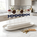 Six assorted red velvet, chocolate, and vanilla cake pops tied with navy ribbon on a ceramic tray beside house keys on white marble.