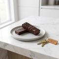 Two Dubai-style chocolate bars with gold foil flecks on a ceramic plate beside a brass key on a white marble counter.