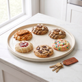 Six assorted artisan welcome-home cookies on a ceramic plate beside brass house keys with a leather welcome-home tag on a white marble counter.